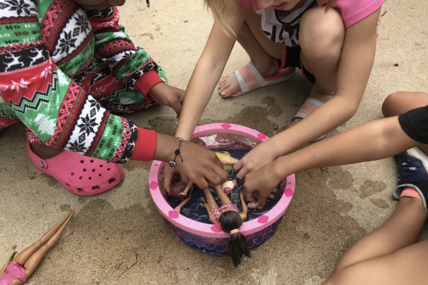 Children play with dolls in a pink basin of water outdoors.